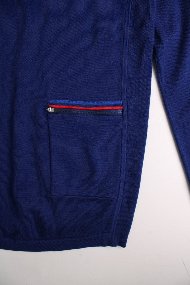 Fred Perry X Bradley Wiggins merino knit jumper in a blue colourway. The Fred Perry logo is embroidered on the front. The knit has side panels, and a small zipped back pocket similar to a cycling jersey. Slim fit silhouette. 