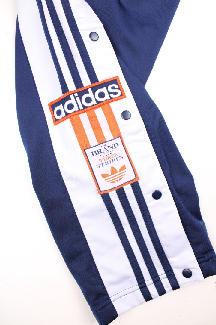 Vintage  Popper Tracksuit Bottoms in a  colourway with stripes going down the sides, has an elasticated waist with drawstring, zip up side pockets, and the logo embroidered on the down the legs. 