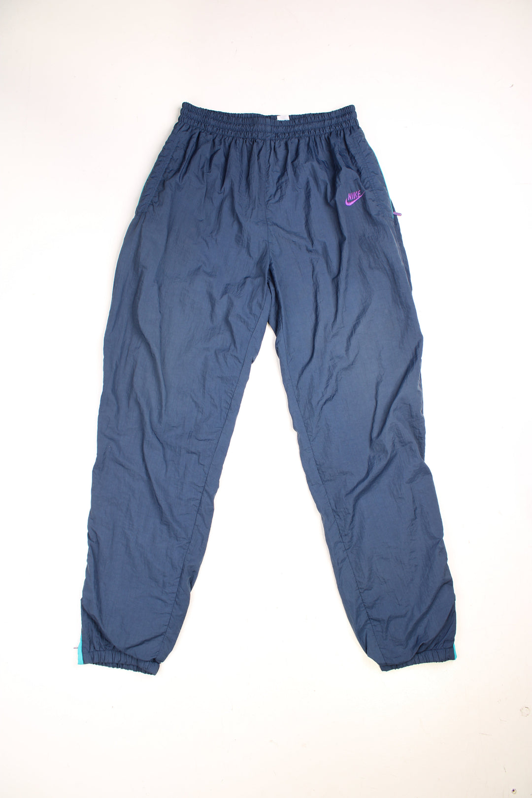 Vintage Nike Shell Tracksuit Bottoms in a blue colourway, has an elasticated waist with drawstring, zip up side pockets, and the logo embroidered in purple on the front. 