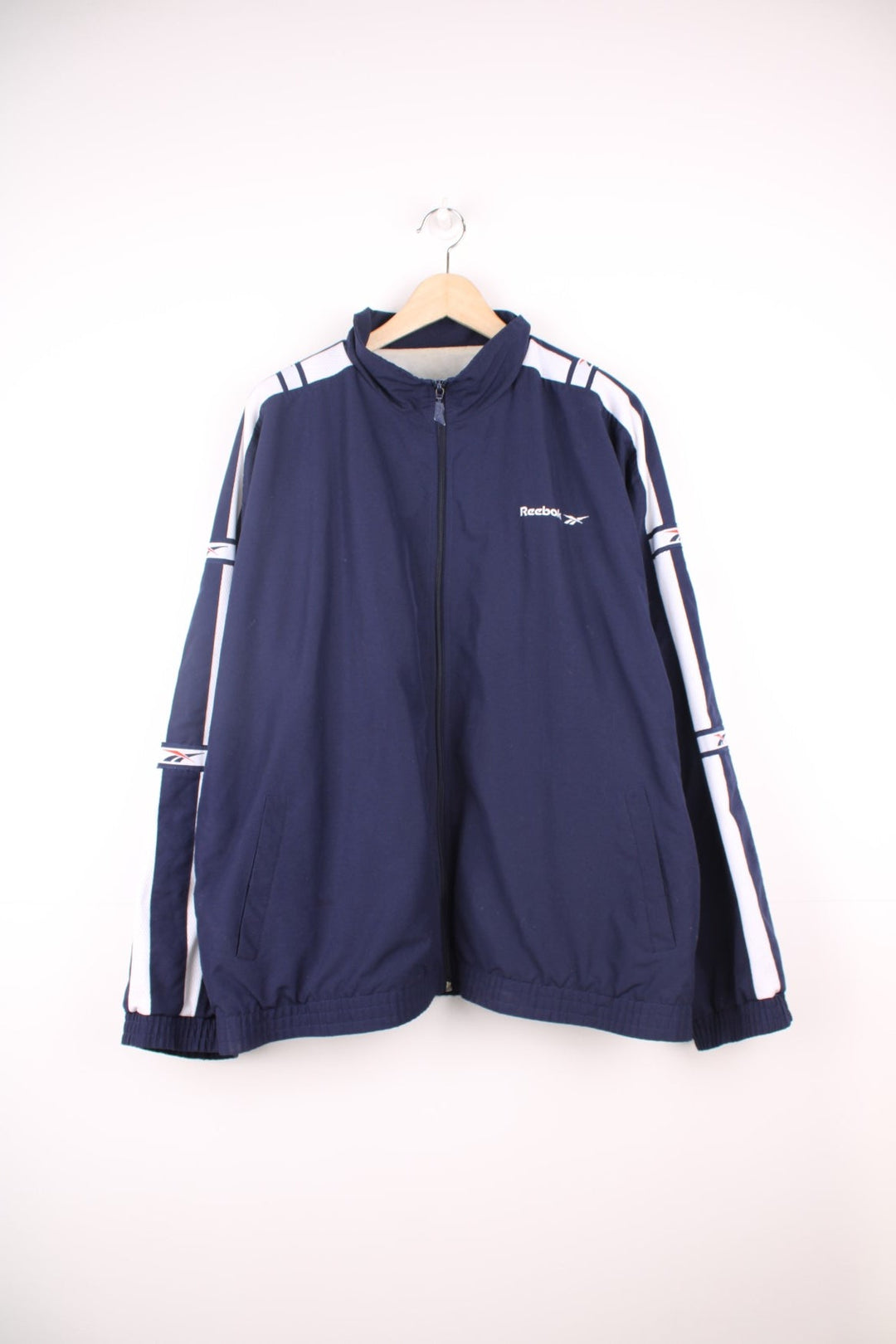 Vintage Reebok tracksuit top in navy blue. Features embroidered logo on the chest, and white tape sleeve with repeated logo.