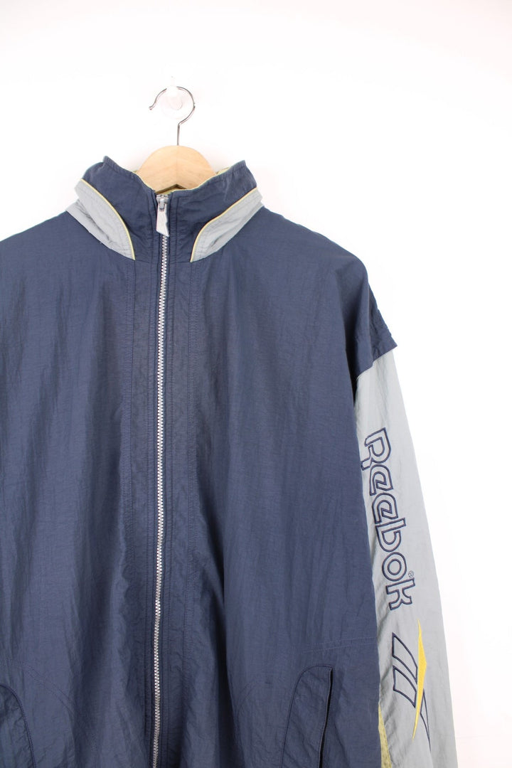 Vintage Reebok zip through tracksuit top in grey and blue. Features embroidered spell out logo down the sleeve, logo on the back, and a pack away hood.  