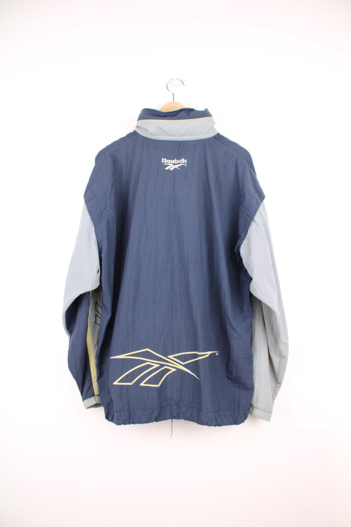 Vintage Reebok zip through tracksuit top in grey and blue. Features embroidered spell out logo down the sleeve, logo on the back, and a pack away hood.  