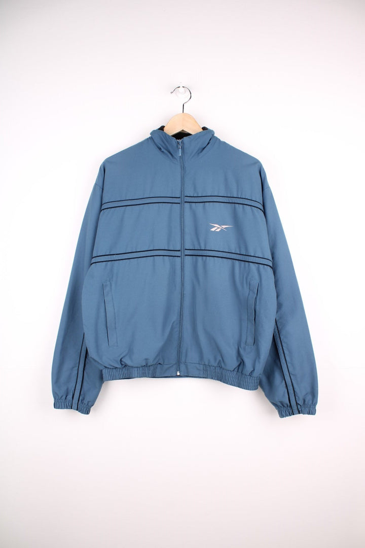 Reebok zip through track jacket in blue with black piping, two pockets, and embroidered logos in white on the front and back. 