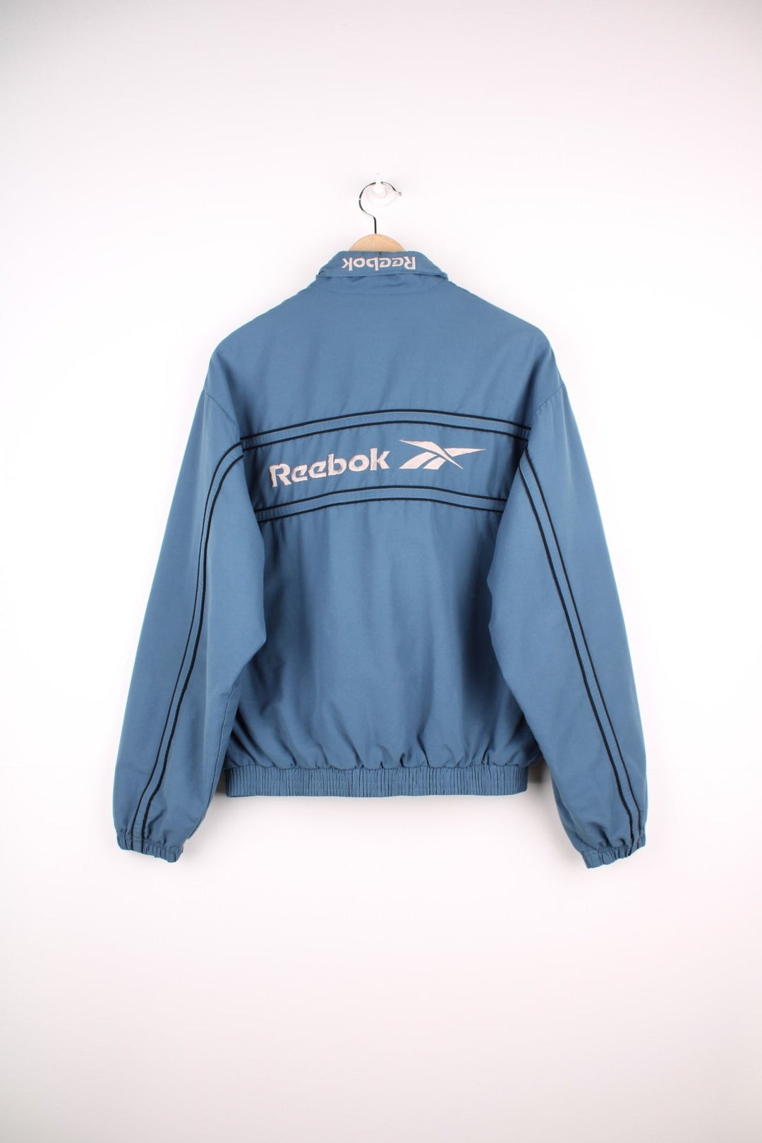 Reebok zip through track jacket in blue with black piping, two pockets, and embroidered logos in white on the front and back. 