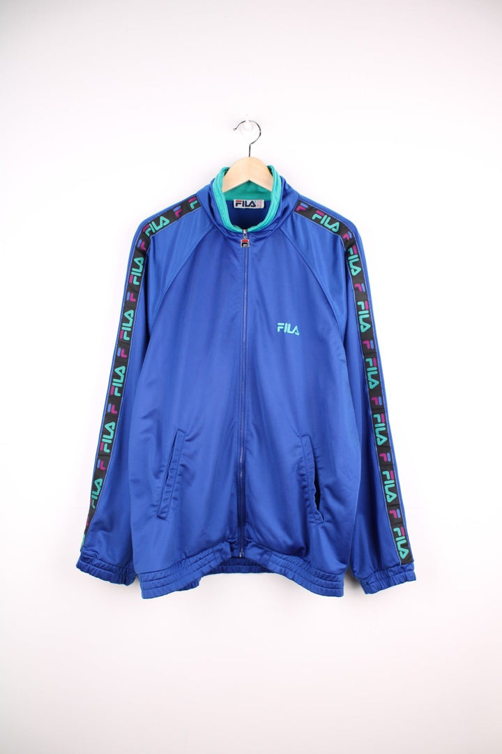 90s Fila tracksuit top in blue with turquoise and fuschia accents, branded ribbon down the sleeves, two pockets, and zip closure. 