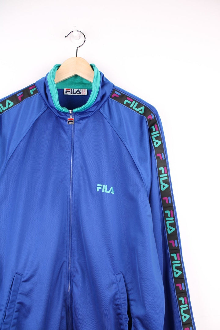 90s Fila tracksuit top in blue with turquoise and fuschia accents, branded ribbon down the sleeves, two pockets, and zip closure. 