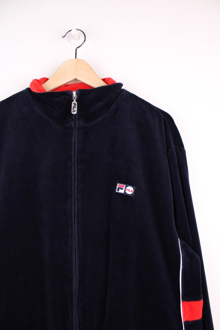 Fila zip-through tracksuit top in navy blue velour with red and white accent stripes on the sleeves, two pockets and an embroidered logo patch on the chest. 