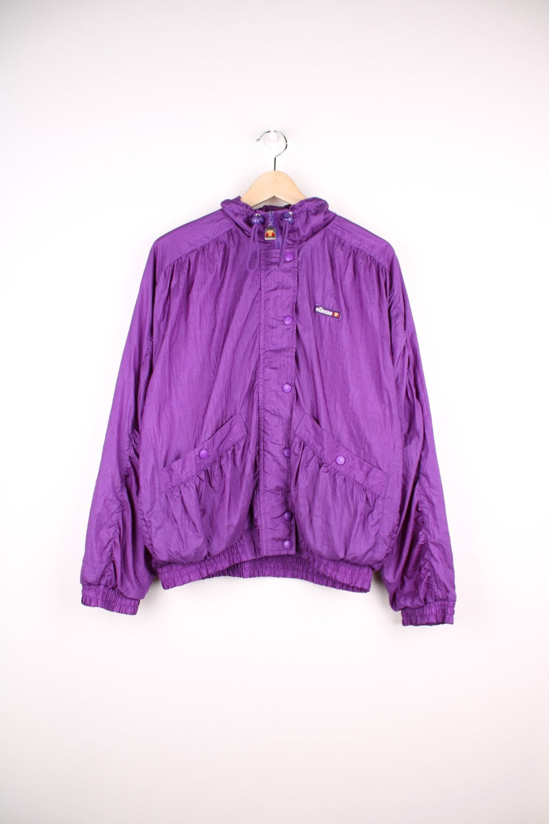 Ellesse track top/shell jacket in bright purple with two pockets, an embroidered logo, snap closure, and toggle adjusters on the collar. 