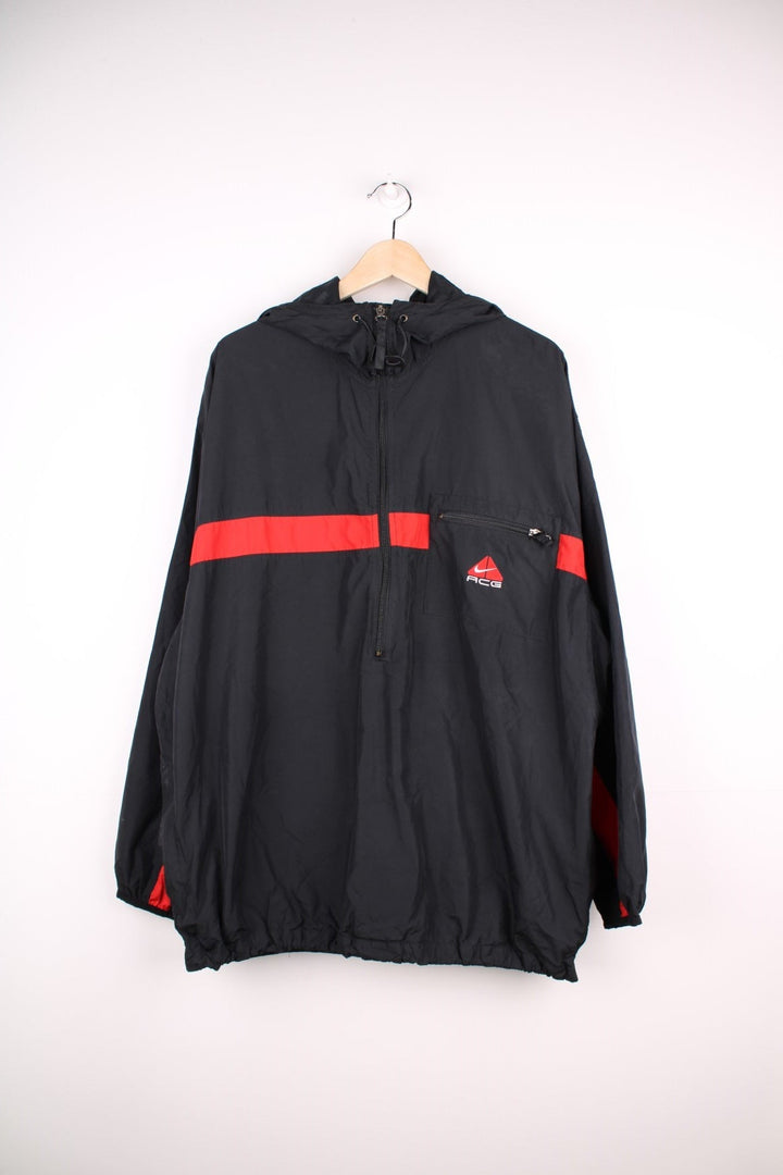 Nike ACG 1/2 zip hooded lightweight windbreaker in black with a central red stripe detail, three zipped pockets, mesh lining, an embroidered ACG logo, and toggle adjusters at the collar. 