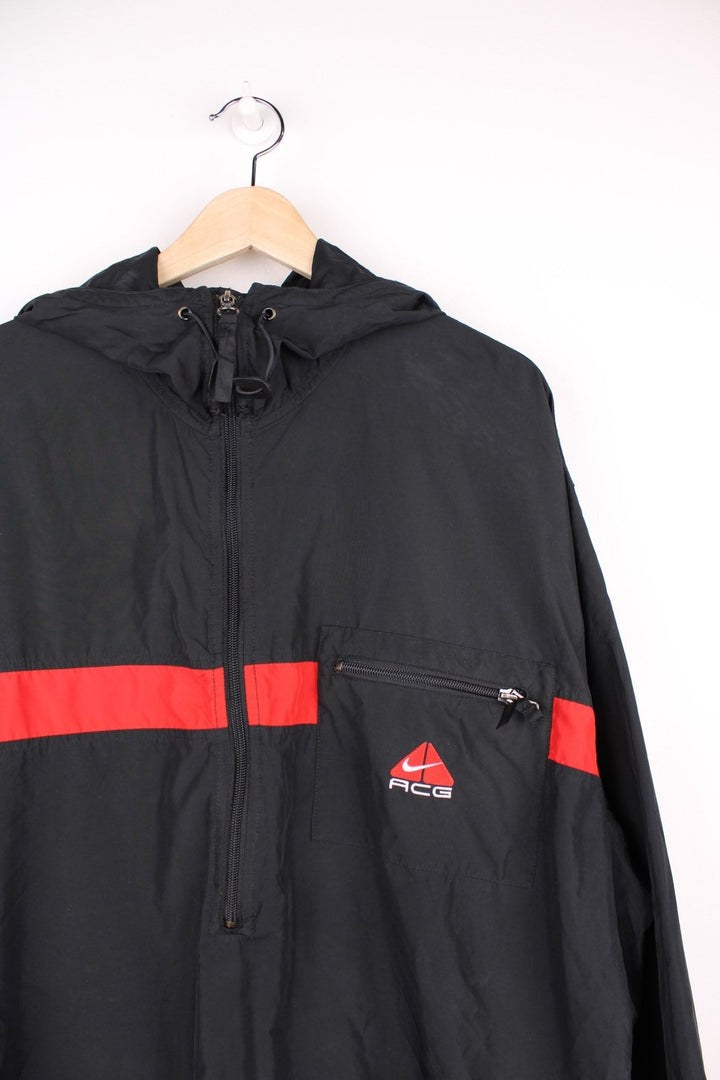 Nike ACG 1/2 zip hooded lightweight windbreaker in black with a central red stripe detail, three zipped pockets, mesh lining, an embroidered ACG logo, and toggle adjusters at the collar. 