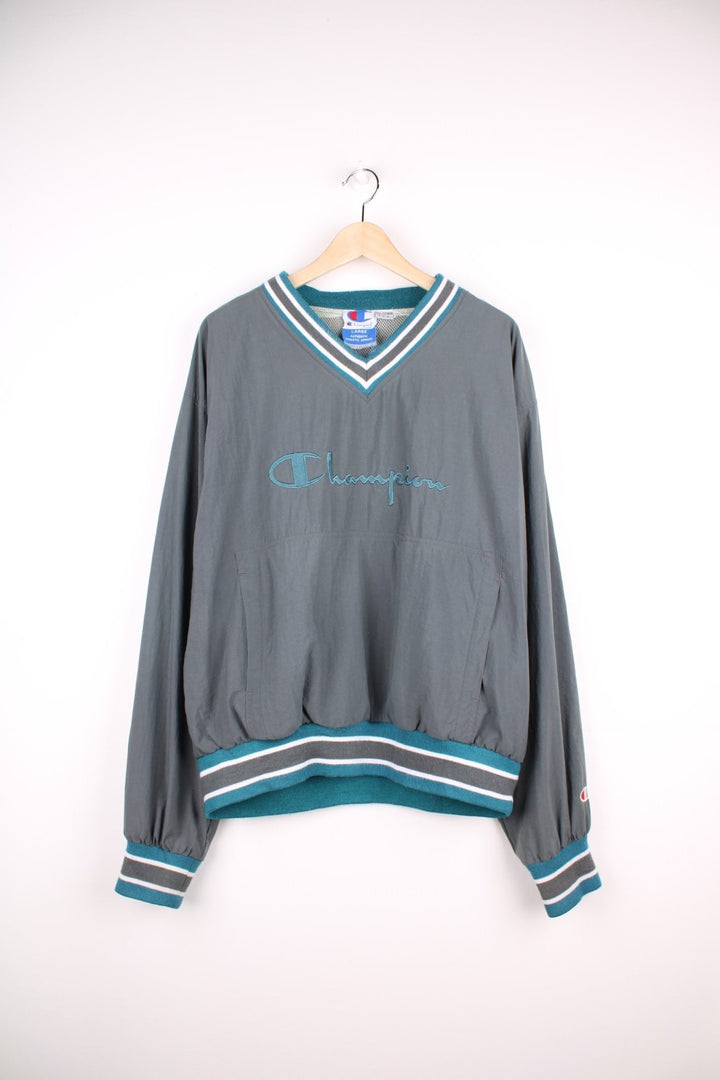 Champion v-neck pullover track top in grey with blue and white striped detailing on hem, cuffs and collar, and embroidered logos on the chest, sleeve and back. 