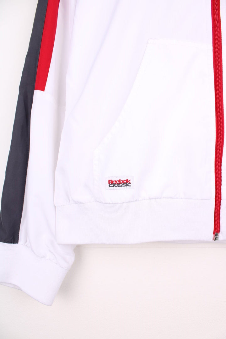 Reebok Classic zip-through tracksuit top in white with red and grey stripes on the sleeves, two pockets and embroidered logos on the chest and back.  