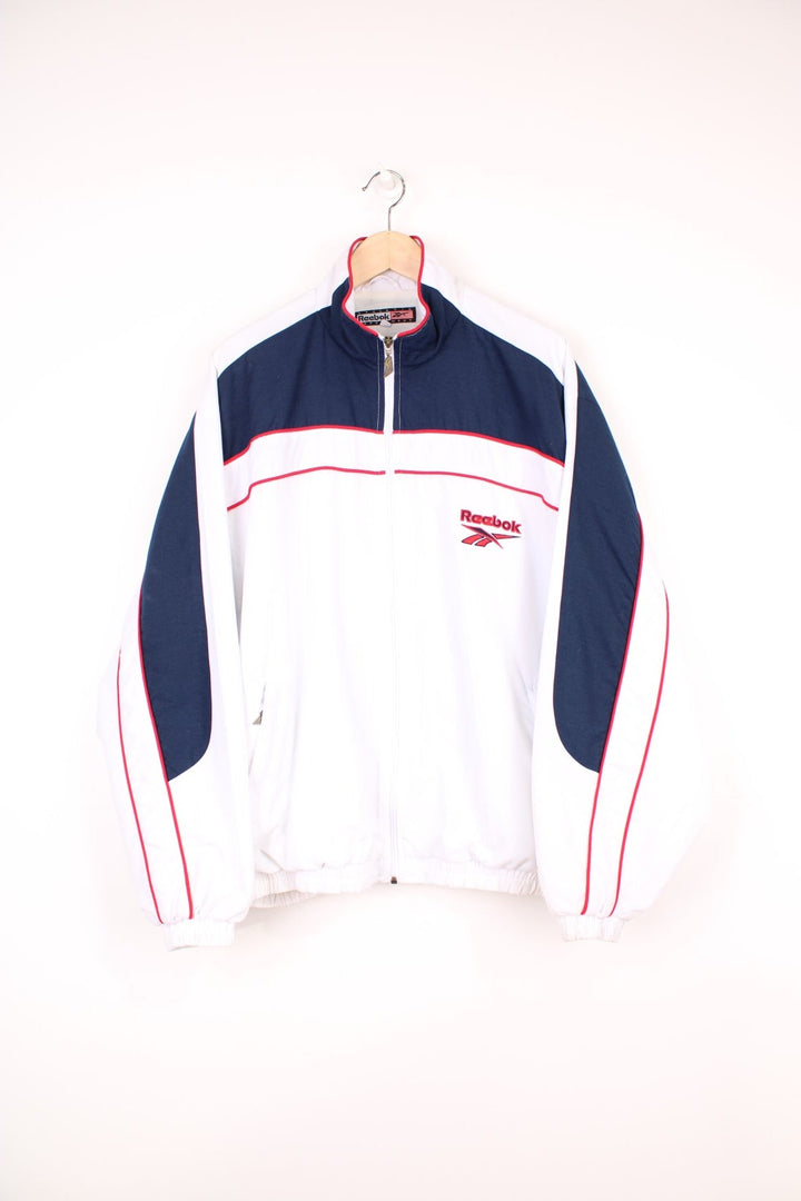 Reebok all white zip through tracksuit top, features embroidered logo on the chest and cuff ,navy blue panels and red piping. 