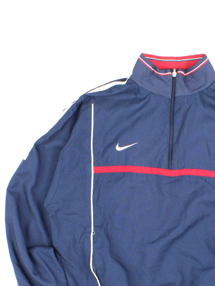 Vintage Nike 1/4 zip reversible pullover track top in navy blue with two zipped pockets, red and white stripe detailing, and a logo on the sleeve. Reverse side is red with blue and white stripe detailing. Swoosh embroidered on the chest of both sides.