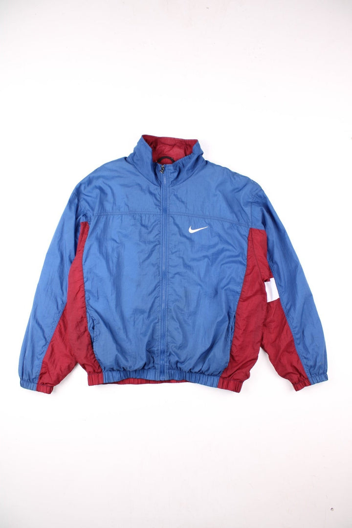 Nike track jacket in blue with red and white panelling, two pockets, zip closure, and an embroidered swoosh logo on the front and back.