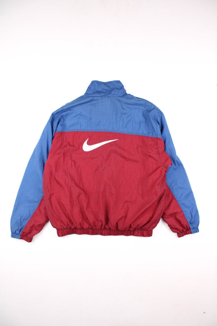 Nike track jacket in blue with red and white panelling, two pockets, zip closure, and an embroidered swoosh logo on the front and back.