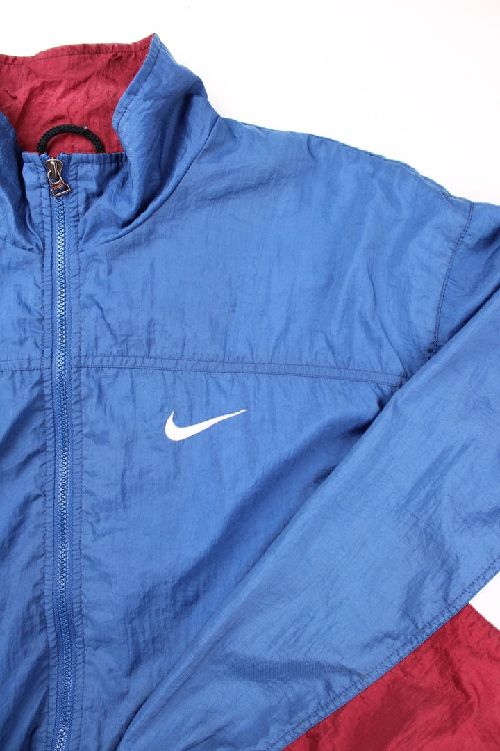 Nike track jacket in blue with red and white panelling, two pockets, zip closure, and an embroidered swoosh logo on the front and back.