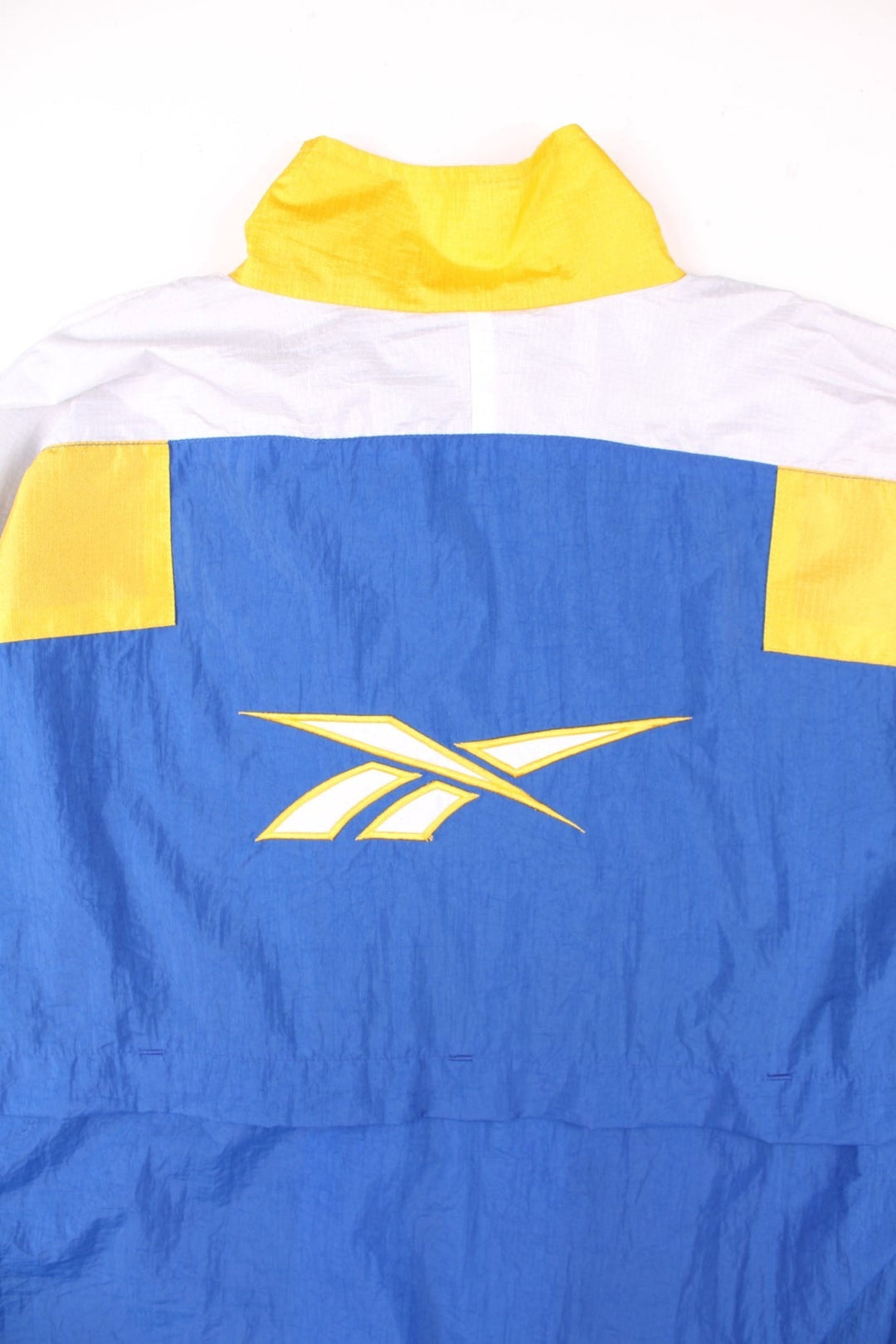 Vintage 90's Reebok Windbreaker Jacket in blue, with yellow and white panelling, two pockets, zip and velcro closure, a drawstring at the hem, and an embroidered logo on the chest and back.