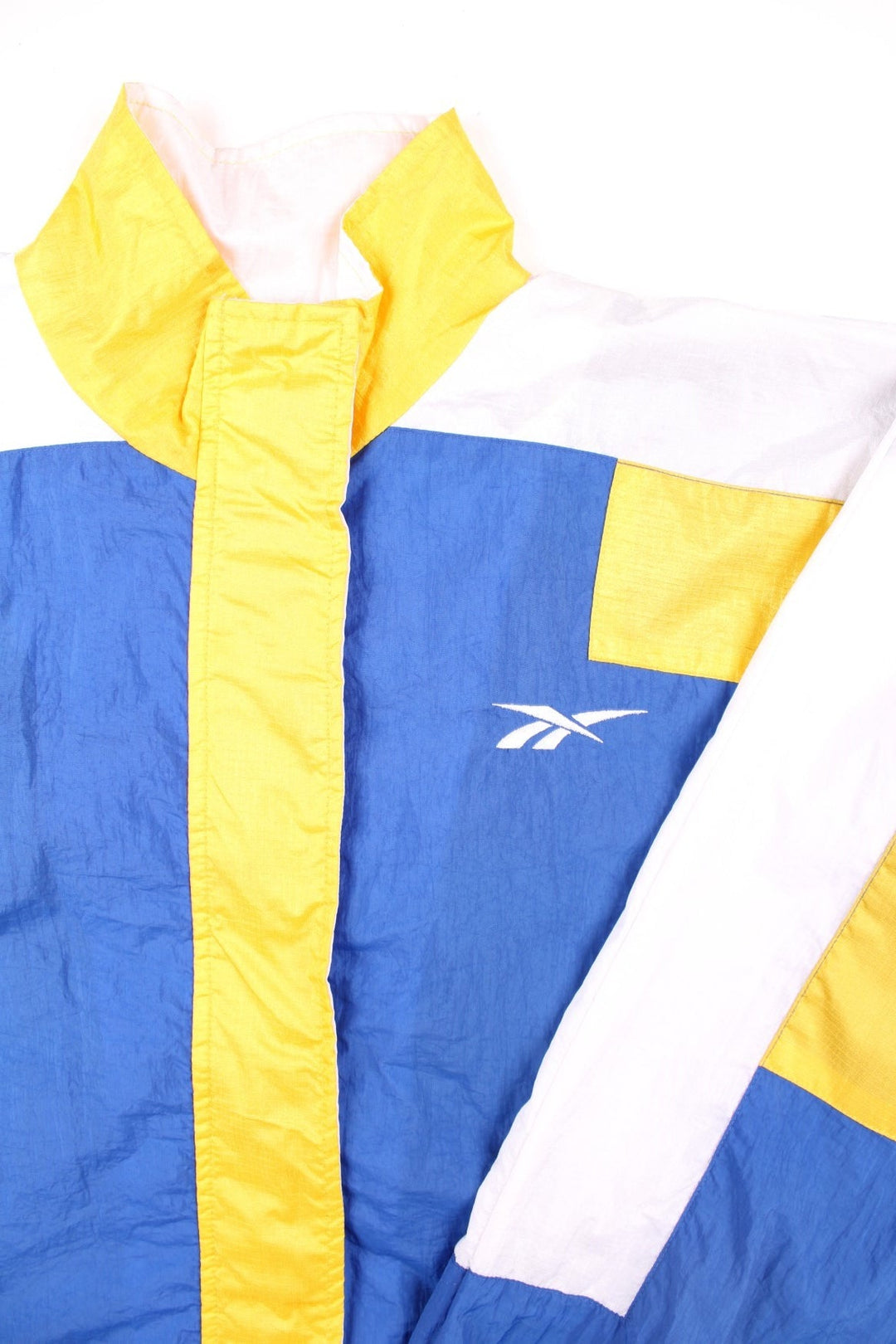 Vintage 90's Reebok Windbreaker Jacket in blue, with yellow and white panelling, two pockets, zip and velcro closure, a drawstring at the hem, and an embroidered logo on the chest and back.