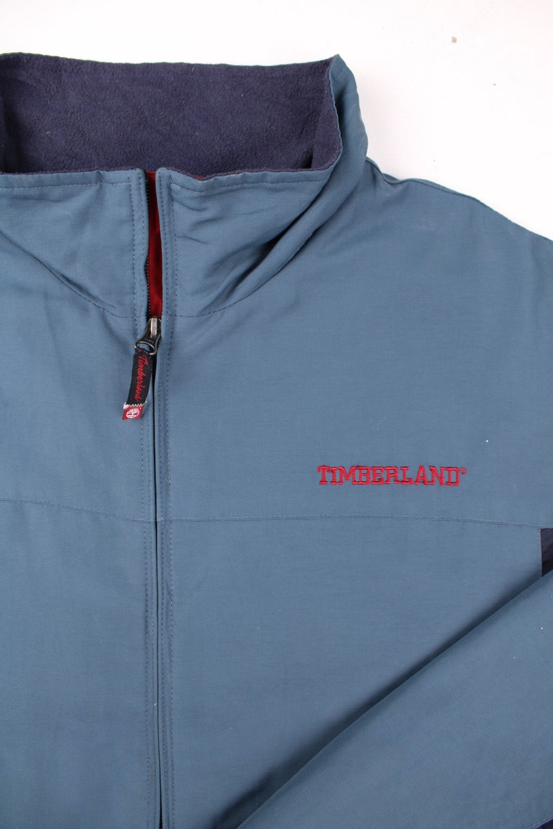 Timberland light jacket in blue-grey, with navy blue panelling, two pockets, zip closure, and an embroidered logo on the chest and back.