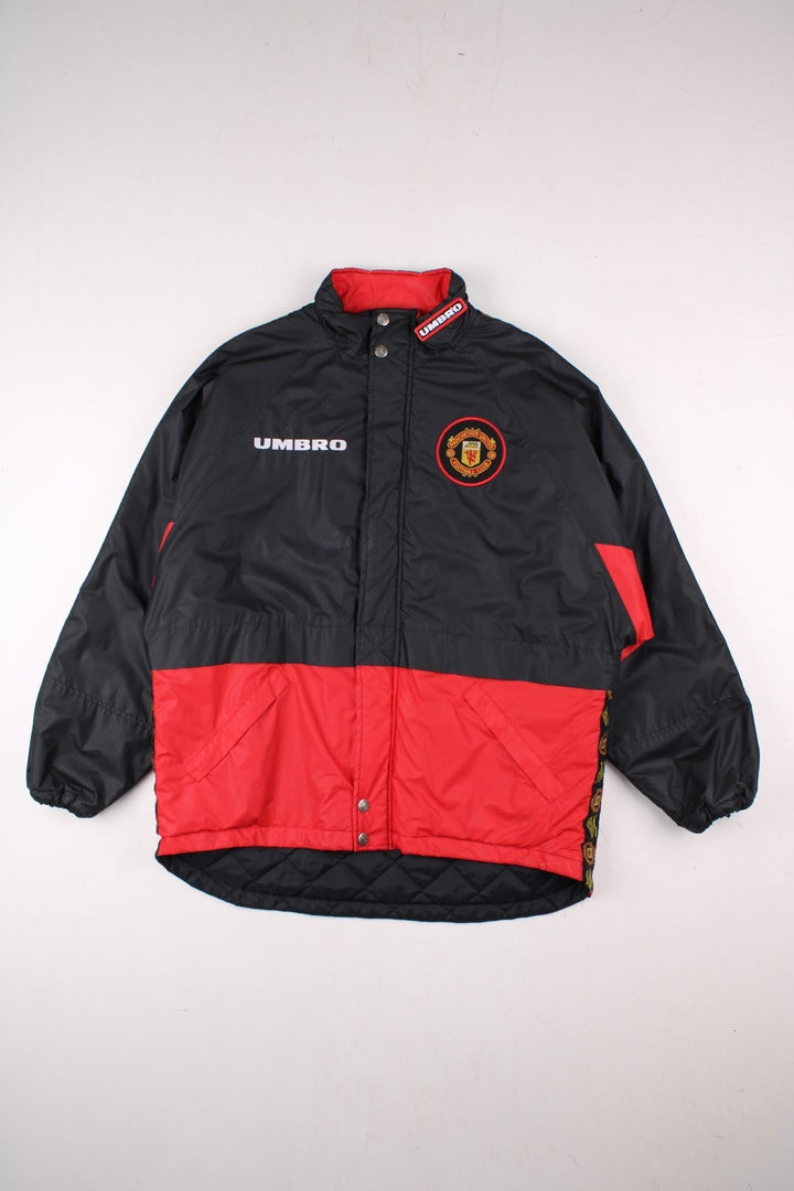 Vintage 1996/97 Manchester United Umbro Coat in black with red panelling, quilt lining, two zipped pockets, zip and snap closure, an embroidered logo on the chest and on the back big logos printed, also has logo tape down each sleeve.