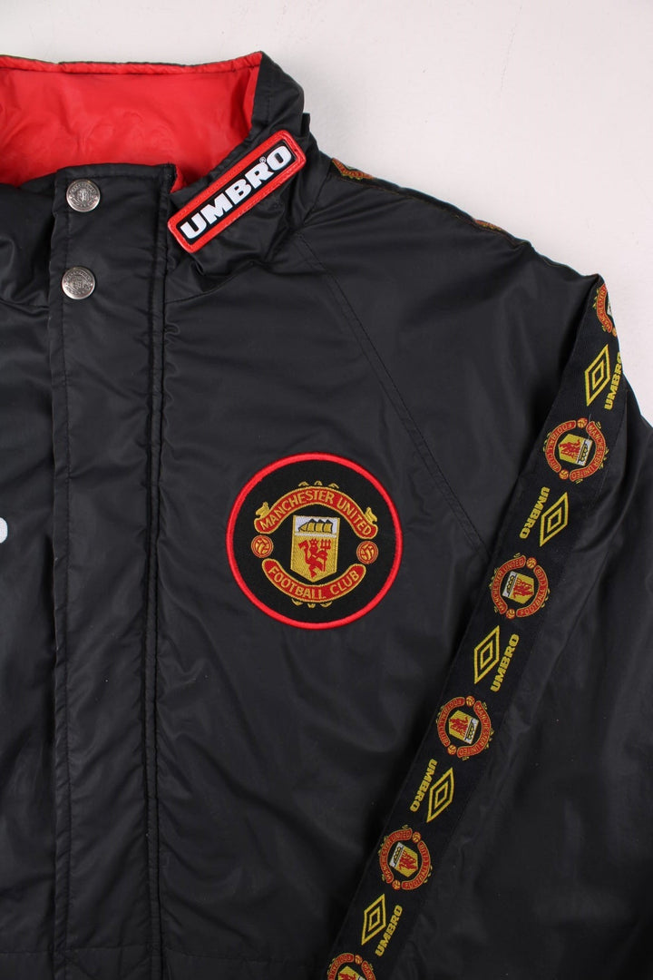 Vintage 1996/97 Manchester United Umbro Coat in black with red panelling, quilt lining, two zipped pockets, zip and snap closure, an embroidered logo on the chest and on the back big logos printed, also has logo tape down each sleeve.