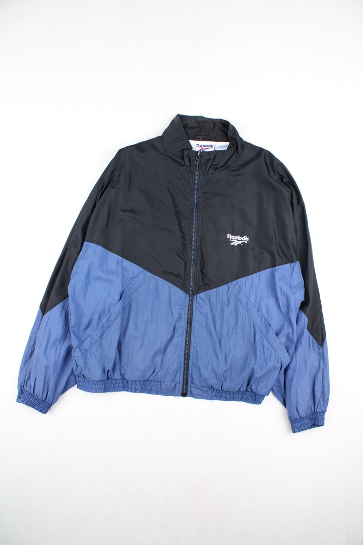 Vintage Reebok Track Top in a black colourway with blue bottom half panelling. Full zip with two side pockets and large reebok logo on the back.