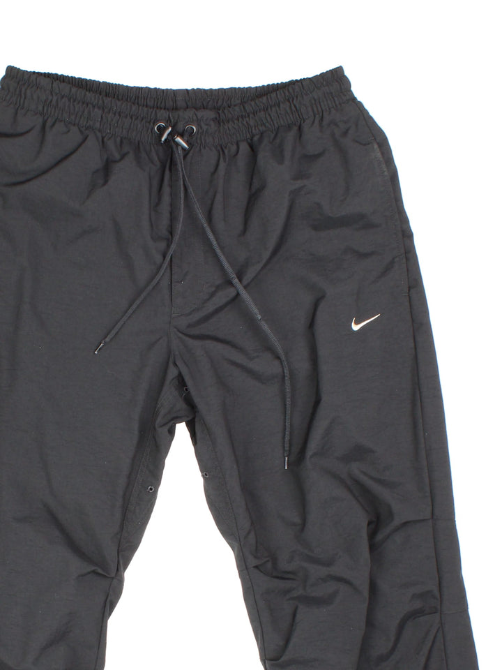 Nike tracksuit bottoms in black with four pockets, a drawstring at the waist, snap-open cuffs and an embroidered swoosh in white.