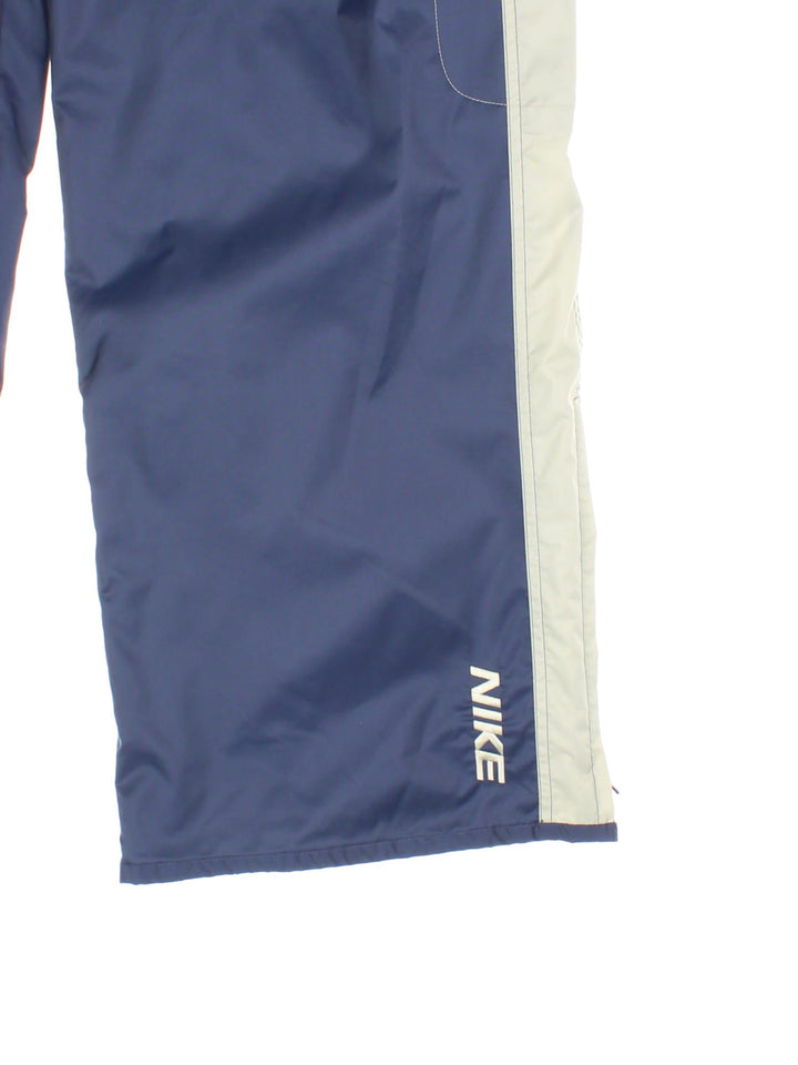 Nike tracksuit bottoms in navy blue with grey stripe detailing, multiple pockets, white contrast stitching, embroidered logos and zip, snap and button closure.