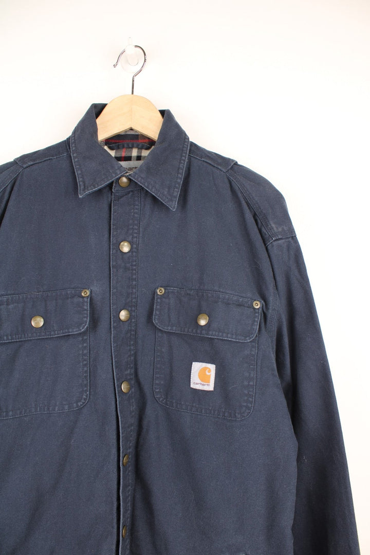 Carhartt Shirt Jacket in a navy blue colourway, heavy cotton material, button up with multiple pockets, has a tartan lining, and the logo embroidered on the front. 