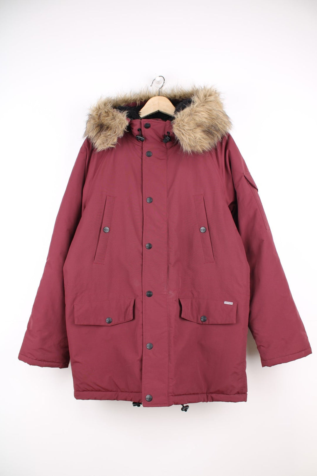 Carhartt Parka Coat in a burgundy colourway, insulated with a quilted lining, has multiple pockets, detachable faux fur hood, and has the logo embroidered on the front and left sleeve.