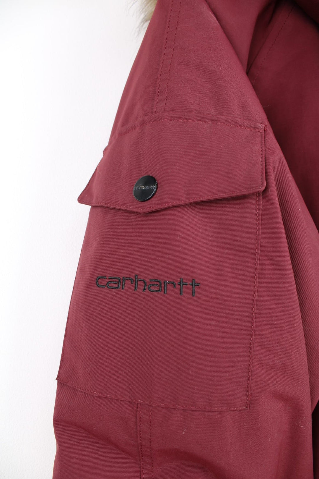 Carhartt Parka Coat in a burgundy colourway, insulated with a quilted lining, has multiple pockets, detachable faux fur hood, and has the logo embroidered on the front and left sleeve.