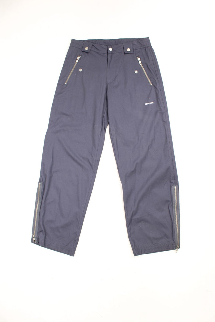 Grey Reebok cotton trousers with zip and popper fastening, zip pockets, zips up both legs and embroidered spell out logo. 