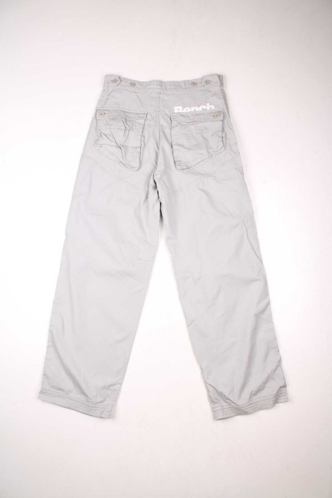 Bench pleated knee cargo pants in tan with four pockets, buttoned adjusters at the waist and printed logos in white above the front left and back right pockets.