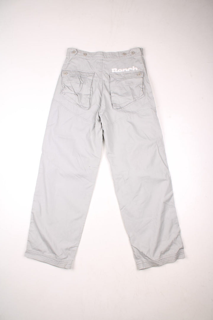 Bench pleated knee cargo pants in tan with four pockets, buttoned adjusters at the waist and printed logos in white above the front left and back right pockets.