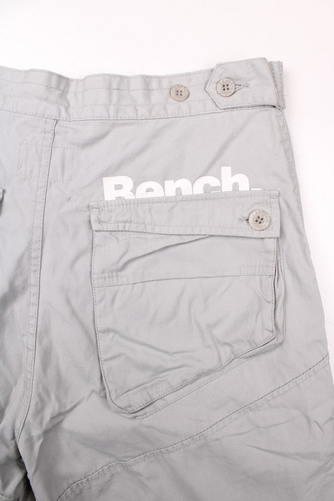 Bench pleated knee cargo pants in tan with four pockets, buttoned adjusters at the waist and printed logos in white above the front left and back right pockets.